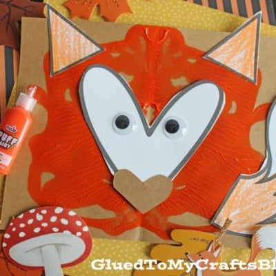 A fox craft made from orange paint, paper, googly eyes, and a heart-shaped nose, with fall-themed decorations and a bottle of orange puff paint on a dark background.