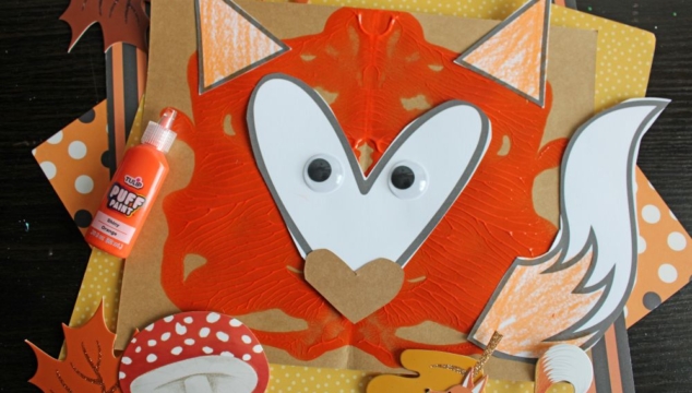 A fox craft made from orange paint, paper, googly eyes, and a heart-shaped nose, with fall-themed decorations and a bottle of orange puff paint on a dark background.