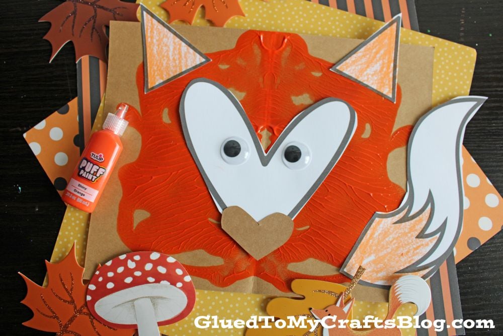 A fox craft made from orange paint, paper, googly eyes, and a heart-shaped nose, with fall-themed decorations and a bottle of orange puff paint on a dark background.