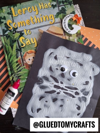 Childrens book Leroy Has Something to Say with an orange paper, white puffy paint, a bear face craft, googly eyes, a glue tube, and an orange bookmark shaped like a fox on a table—perfect inspiration for creating your own paint splat ghost project.