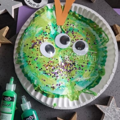 A paper plate painted green with googly eyes, orange paper antennae, and glitter, resembling a caterpillar face. Craft supplies and stars surround the plate. @GLUEDTOMYCRAFTS is labeled at the bottom.