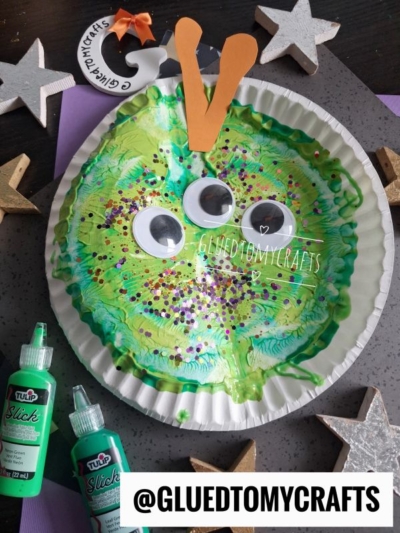 A paper plate painted green with googly eyes, orange paper antennae, and glitter, resembling a caterpillar face. Craft supplies and stars surround the plate. @GLUEDTOMYCRAFTS is labeled at the bottom.