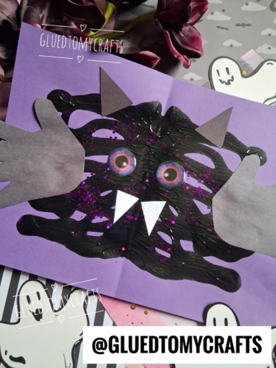 Childrens craft of a black paint splat bat monster with googly eyes, gray hand shapes for arms, and white triangle fangs on purple paper. Halloween-themed background with ghosts. @GLUEDTOMYCRAFTS watermark.
