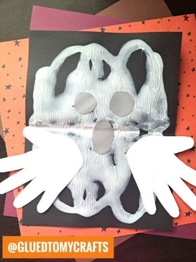 A ghost craft made from white paint on black paper, with cut-out gray eyes and mouth, plus handprint cut-outs for wings. Orange star-patterned paper is underneath.