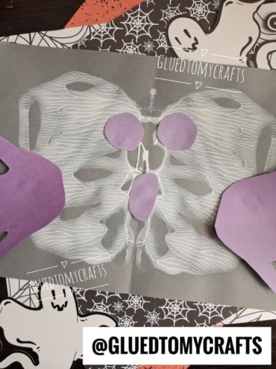 A paper craft of a white skeleton on black paper, with purple paper hands and circles, sits on a Halloween-themed background with ghosts, webs, bats, and a paint splat ghost. @GLUEDTOMYCRAFTS is written in white text.