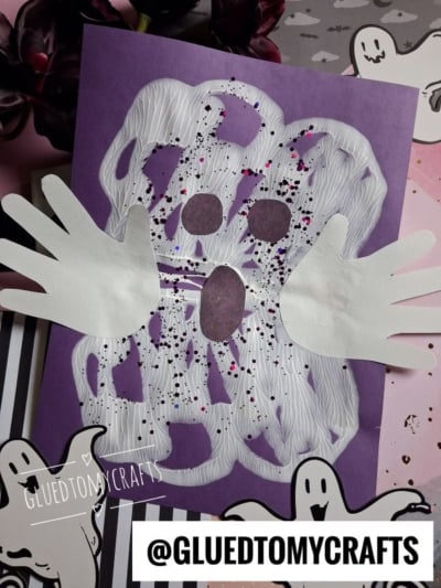 A paint splat ghost craft made from white paint handprints on purple paper, decorated with two paper hands, black oval eyes and mouth, and sprinkled with black and pink glitter. Cartoon ghost stickers and @GLUEDTOMYCRAFTS text are visible.