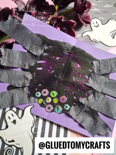 A playful paint splat spider craft for children, featuring black paint splat bodies, tissue paper legs, and colorful googly eyes on vibrant purple paper. The artwork is adorned with ghost cutouts and surrounded by flowers, while the @GLUEDTOMYCRAFTS text is visible.