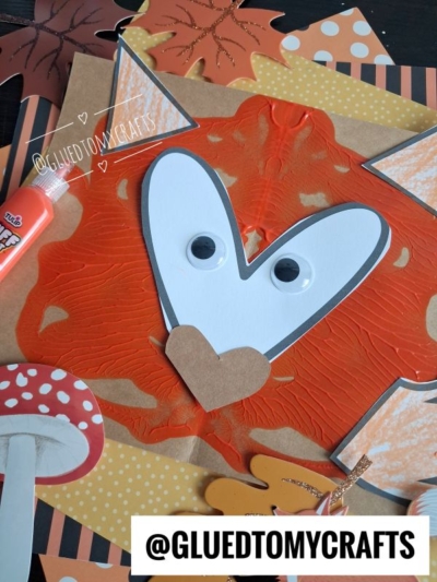 A fox face made from orange paint, googly eyes, and paper, surrounded by autumn-themed paper cutouts, glue, and a mushroom decoration. This charming PAINT SPLAT CRAFTS project features the handle @GLUEDTOMYCRAFTS at the bottom.