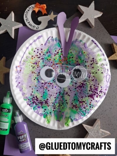 A colorful paper plate butterfly craft with splattered paint, large googly eyes, purple antennae, and glitter. Puff paint bottles and silver star decorations are nearby. The handle @GLUEDTOMYCRAFTS is at the bottom.