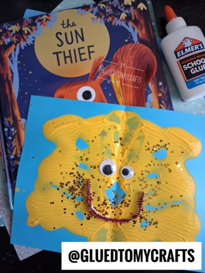 A childrens book titled The Sun Thief, a bottle of Elmer’s glue, and a yellow sun-shaped craft with googly eyes, glitter, and a smile are shown on a table—perfect inspiration for summer craft ideas. @GLUEDTOMYCRAFTS is written at the bottom.