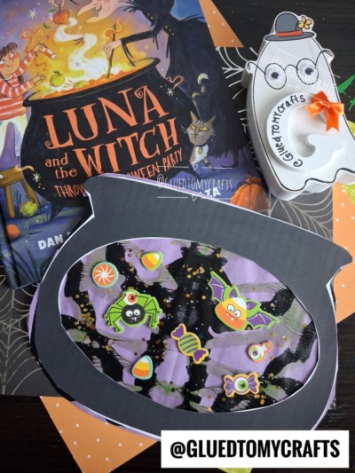 A witch-themed children’s book perfect for halloween story time, Halloween crafts including a decorated cauldron cutout with stickers, and a ghost-shaped treat box with glasses and an orange ribbon bow, arranged on a table. @GLUEDTOMYCRAFTS text at the bottom.