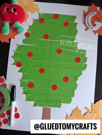 A craft tree made from green painter’s tape strips with red dot stickers as apples on white paper, surrounded by a toy apple, artificial leaves, marker, and crafting supplies—an adorable example of painter’s tape craft ideas.