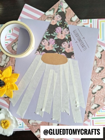 A gnome craft with a floral hat, brown nose, and white tape beard on purple paper. Surrounded by washi tape, felt flowers, and pastel-patterned papers. @GLUEDTOMYCRAFTS label is at the bottom.