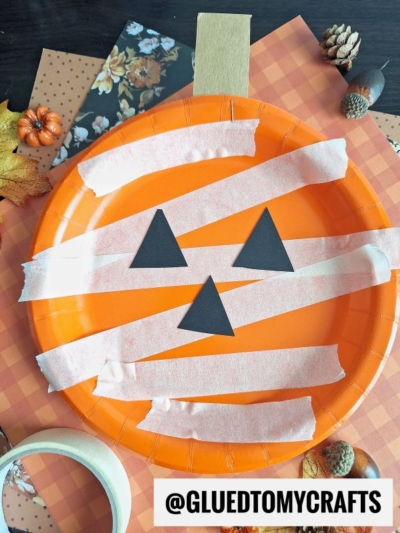 Orange paper plate pumpkin with black triangle eyes and nose, covered in white paper strips like a mummy, on a fall-themed background with leaves and small pumpkins. Text reads @GLUEDTOMYCRAFTS.