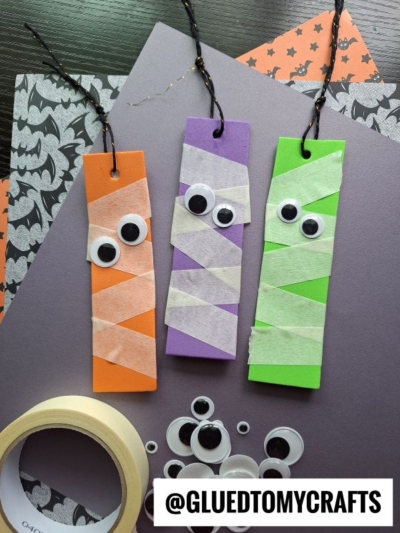 Three rectangular bookmarks in orange, purple, and green are decorated like mummies with googly eyes and white tape strips. Craft supplies like tape and eyes are nearby. The @GLUETOMYCRAFTS handle is shown at the bottom.