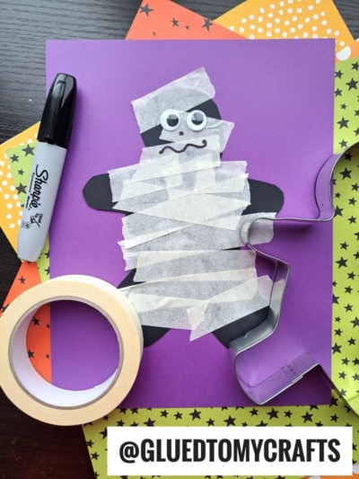 A paper gingerbread person is mummified with tape on purple paper, next to a marker, tape roll, star papers, and a cookie cutter. Text reads @GLUEDTOMYCRAFTS..