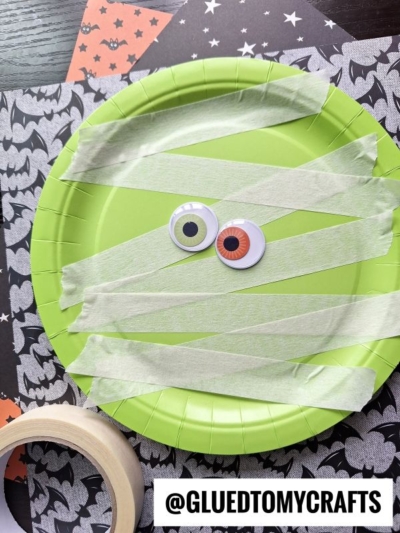 A green paper plate with googly eyes and tape strips to look like a mummy, placed on bat-patterned paper. A roll of masking tape and the text @GLUEDTOMYCRAFTS are also visible.