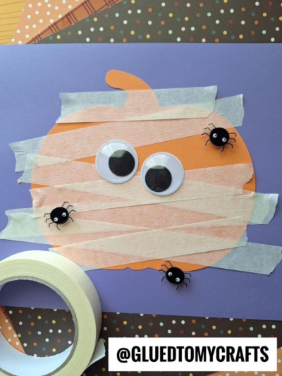 A paper pumpkin craft with googly eyes, tape bandages, and black button spiders on a purple background. A roll of tape and patterned paper are nearby.
