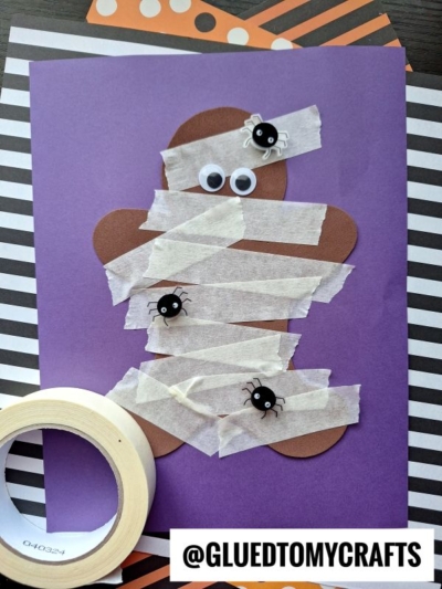 A gingerbread-shaped paper figure on purple paper, wrapped with white tape to look like a mummy. Googly eyes and plastic spiders are attached. Tape and striped paper are in the background. @GLUEDTOMYCRAFTS is at the bottom.