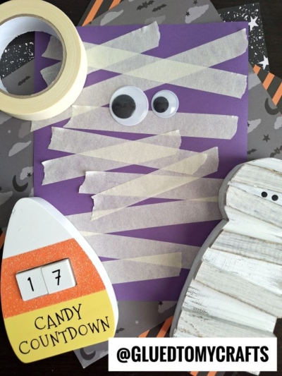Purple paper with strips of masking tape arranged like a mummy, two googly eyes, a roll of tape, a candy countdown block showing 17, and a wooden ghost on a Halloween-themed background.