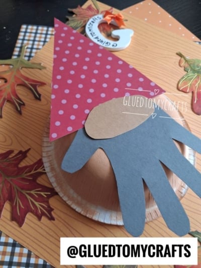 Paper plate gnome craft with a red polka-dot hat and a handprint beard on an autumn-themed background, featuring colorful paper leaves and an @GLUEDTOMYCRAFTS tag.