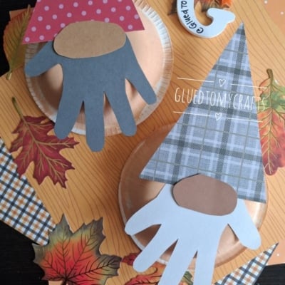 Two gnome crafts made with paper plates, cut-out hand shapes as beards, and patterned paper hats. They are surrounded by paper autumn leaves on an orange background. @GLUEDTOMYCRAFTS is written on the image.