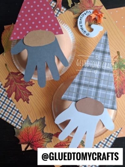 Two gnome crafts made with paper plates, cut-out hand shapes as beards, and patterned paper hats. They are surrounded by paper autumn leaves on an orange background. @GLUEDTOMYCRAFTS is written on the image.
