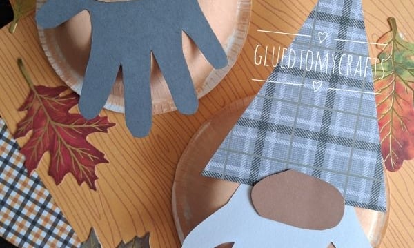 Two gnome crafts made with paper plates, cut-out hand shapes as beards, and patterned paper hats. They are surrounded by paper autumn leaves on an orange background. @GLUEDTOMYCRAFTS is written on the image.