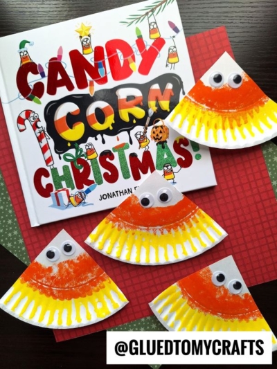Four paper plate candy corn crafts with googly eyes surround the book Candy Corn Christmas on a red and green background. These adorable paper plate candy corn creations add a playful touch, while a GLUEDTOMYCRAFTS label is at the bottom.