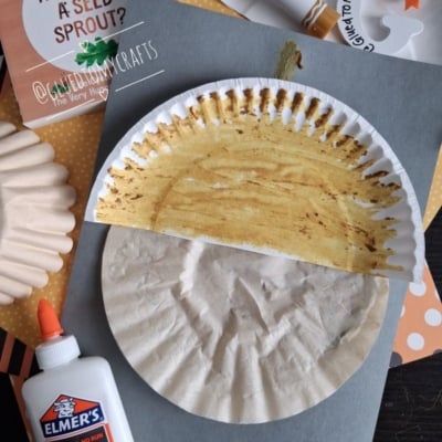 Paper plate craft resembling an acorn, surrounded by glue, craft supplies, and a How Does a Seed Sprout? booklet on a dark surface with an @GLUEDTOMYCRAFTS label at the bottom.
