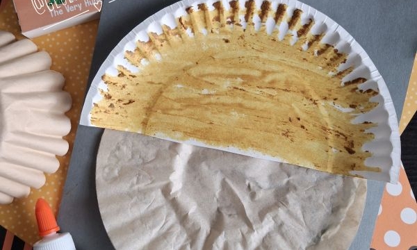 Paper plate craft resembling an acorn, surrounded by glue, craft supplies, and a How Does a Seed Sprout? booklet on a dark surface with an @GLUEDTOMYCRAFTS label at the bottom.