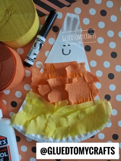 A paper candy corn craft with torn tissue paper in yellow, orange, and white sections, a smiley face, crepe paper rolls, a glue bottle, and a black marker on a polka dot background. Text: @GLUEDTOMYCRAFTS.