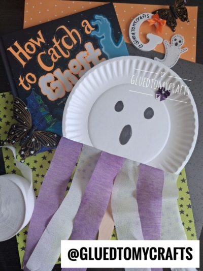 A paper plate ghost craft with purple and white streamers, black eyes and mouth, and a bow sits on a table with the book How to Catch a Ghost and butterflies—a perfect addition to your Halloween Crafts for Kids. @gluedtomycrafts tag is on the image.