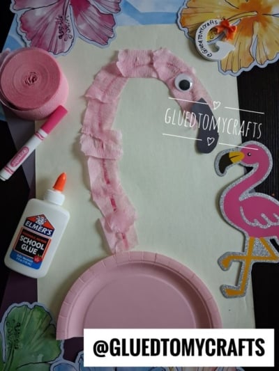 A torn crepe paper flamingo craft made with a pink plate, tissue paper, googly eye, and paper beak. Craft supplies like glue, streamer, and marker are nearby. Decorative hibiscus and a flamingo cutout are in the background.