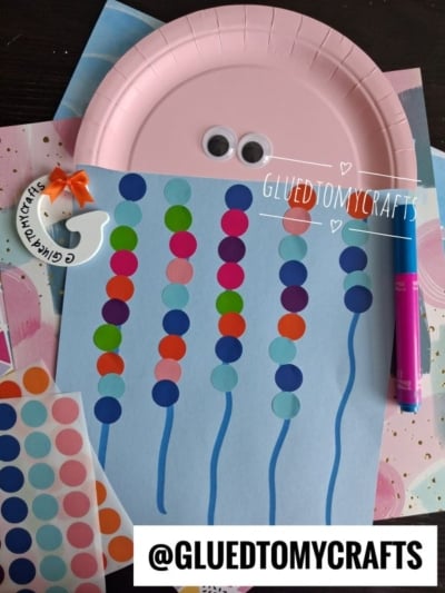 A craft project featuring a blue paper octopus with colorful dot stickers for tentacles, a pink paper plate head adorned with googly eyes, and a pink marker is displayed on a dark surface—an inspiring example of coding label crafts brought to life through creativity and fun materials.