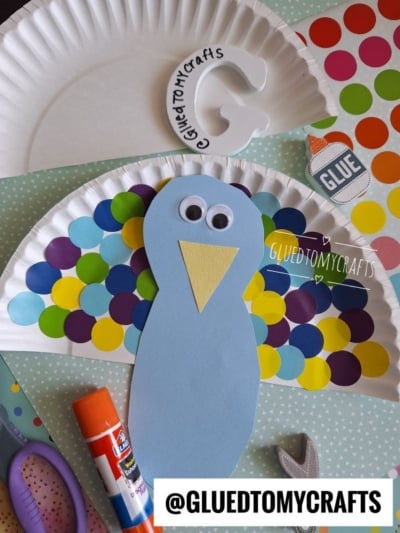 Paper plate peacock craft with colorful paper dots, glue, scissors, and a foam letter C, displayed on polka dot paper. Perfect for kids who enjoy coding label crafts, the peacock features googly eyes and a yellow beak.
