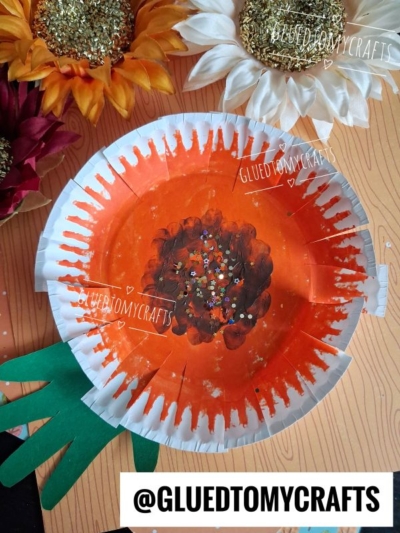 A paper plate painted orange with a brown center and gold glitter, resembling a sunflower, with green paper hand-shaped leaves attached—the perfect idea for FALL CRAFTS FOR KIDS. Decorative sunflowers sit nearby on a wood-textured surface. Text: @GLUEDTOMYCRAFTS.