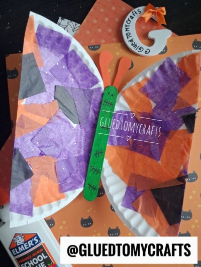 A butterfly craft made from a green popsicle stick and a paper plate with purple, orange, and black tissue paper, on a Halloween-themed background. Perfect for FALL CRAFTS FOR KIDS, Elmers glue and a @GLUEDTOMYCRAFTS tag are visible.