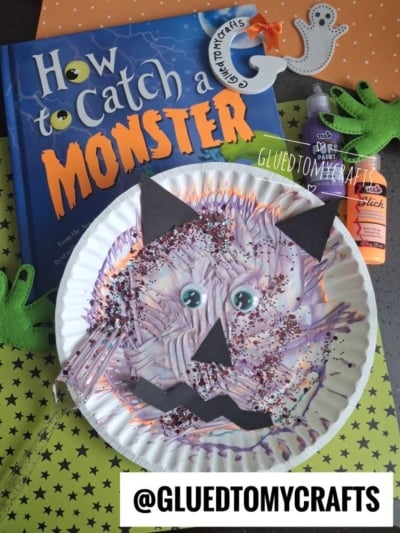 Paper plate monster craft with purple paint and glitter, black paper shapes for facial features, and googly eyes—perfect for Halloween Crafts for Kids. Book titled How to Catch a Monster and Halloween-themed supplies are in the background.