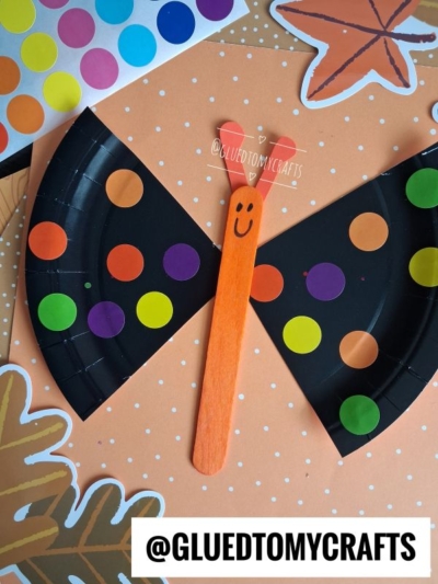 A fun Halloween Crafts for Kids project: a butterfly craft made from a black paper plate, colorful dot stickers, and an orange craft stick with a smiley face, displayed on a polka dot background with leaf and star stickers.