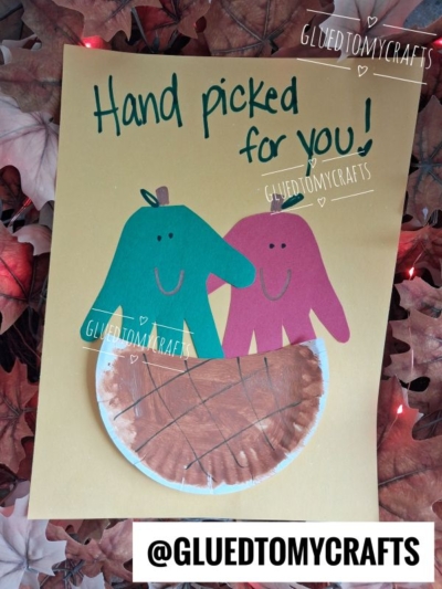 A handmade craft showing two handprint cutouts as people inside a paper plate basket, with the words “Hand picked for you!” on yellow paper and autumn leaves in the background—perfect for those looking for creative FALL CRAFTS FOR KIDS.