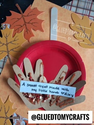 A red paper plate with handprint cutouts as a pie, decorated with glitter, sits on fall leaves. A note reads, A sweet treat made with my little hands ♥ AKY. @GLUEDTOMYCRAFTS is written at the bottom.