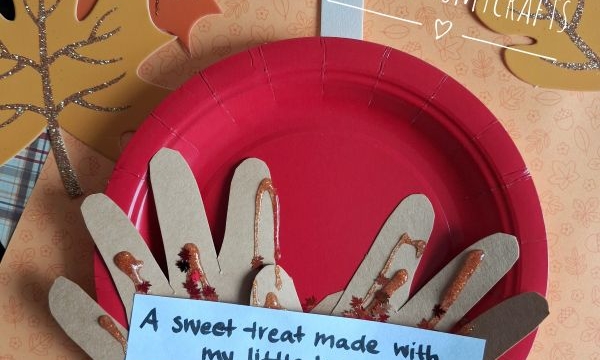 A red paper plate with handprint cutouts as a pie, decorated with glitter, sits on fall leaves. A note reads, A sweet treat made with my little hands ♥ AKY. @GLUEDTOMYCRAFTS is written at the bottom.