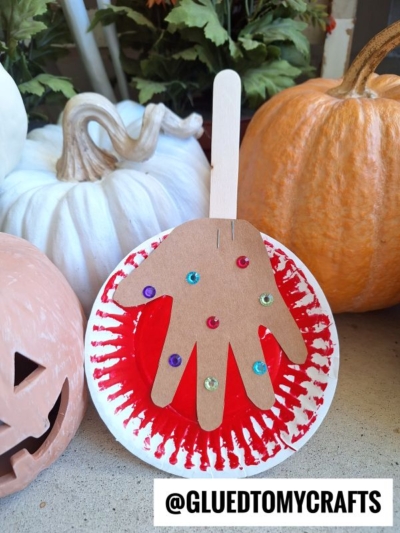A paper plate caramel apple decorated with red paint and sequins, topped with a brown handprint cutout and craft stick, sits among pumpkins. The text @GLUEDTOMYCRAFTS appears in the bottom right corner.