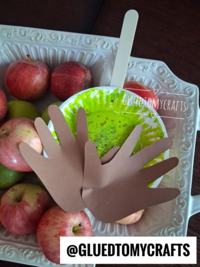 A bowl of green slime with glitter and a popsicle stick is surrounded by apples, next to a paper plate caramel apple craft. Two brown paper hand cutouts rest on top. @GLUEDTOMYCRAFTS is written at the bottom.