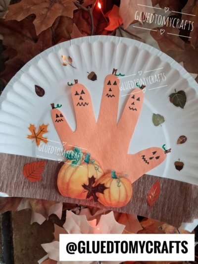 A handprint craft on a paper plate features pumpkins and tiny jack-o-lantern faces on fingers, with autumn leaves as decoration for a festive paper plate pumpkin patch. The scene includes fall colors and a handle @GLUEDTOMYCRAFTS.