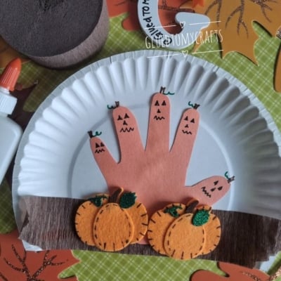 Paper plate craft with a handprint decorated as jack-o-lanterns, felt pumpkins, brown paper for soil, and autumn leaves in the background. Bottle of glue and @GLUEDTOMYCRAFTS text at the bottom.