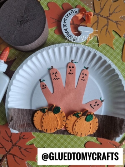 Paper plate craft with a handprint decorated as jack-o-lanterns, felt pumpkins, brown paper for soil, and autumn leaves in the background. Bottle of glue and @GLUEDTOMYCRAFTS text at the bottom.
