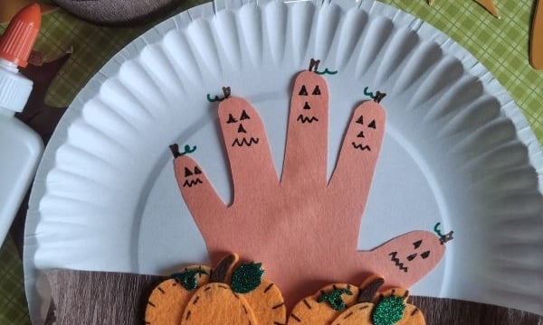 Paper plate craft with a handprint decorated as jack-o-lanterns, felt pumpkins, brown paper for soil, and autumn leaves in the background. Bottle of glue and @GLUEDTOMYCRAFTS text at the bottom.