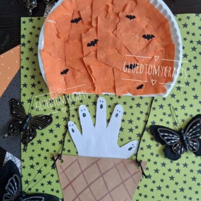 Top-down flat-lay of a kids' Halloween craft with an orange crepe paper pumpkin hot air balloon, handprint ghosts peeking from a paper basket, and decorative black butterflies on a lime-green star-patterned background.